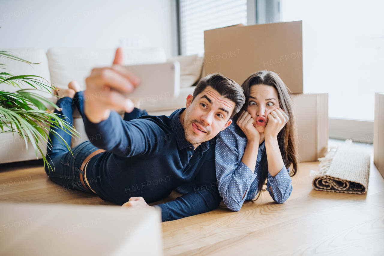 A young happy couple with a smartphone and cardboard boxes lying on a floor, taking selfie and making a face when moving in a new home.