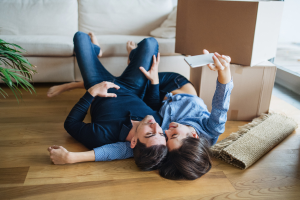 A young happy couple with a smartphone and cardboard boxes lying on a floor, taking selfie when moving in a new home.