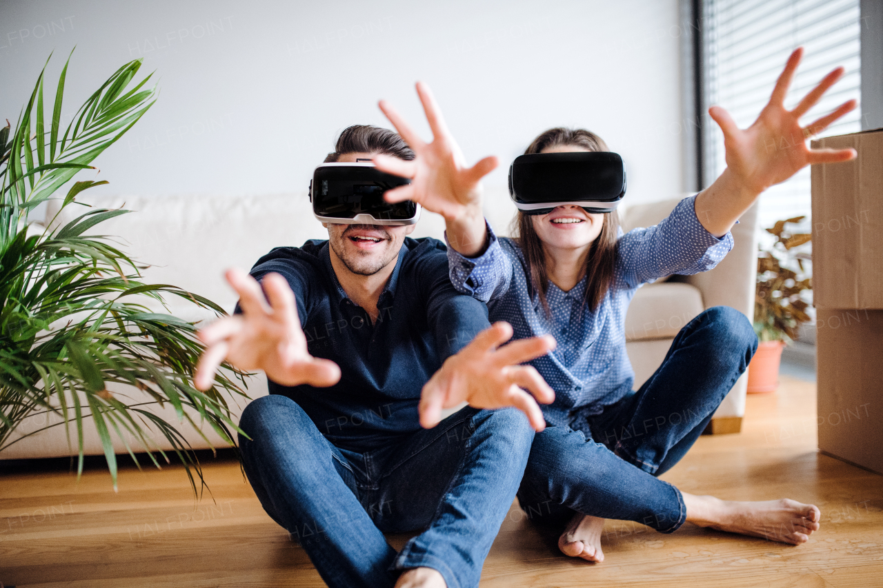 A young couple with VR goggles and cardboard boxes sitting on a floor, moving in a new home.