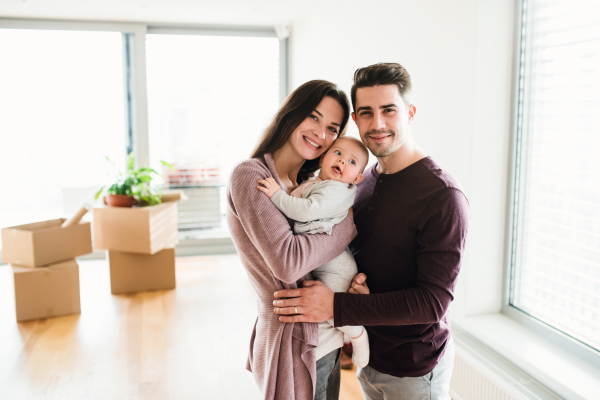 A portrait of happy young couple with a baby and cardboard boxes, moving in a new home.