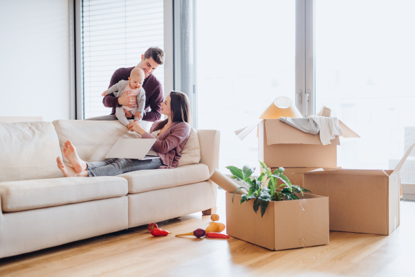 A portrait of happy young couple with a baby,laptop and cardboard boxes, moving in a new home.