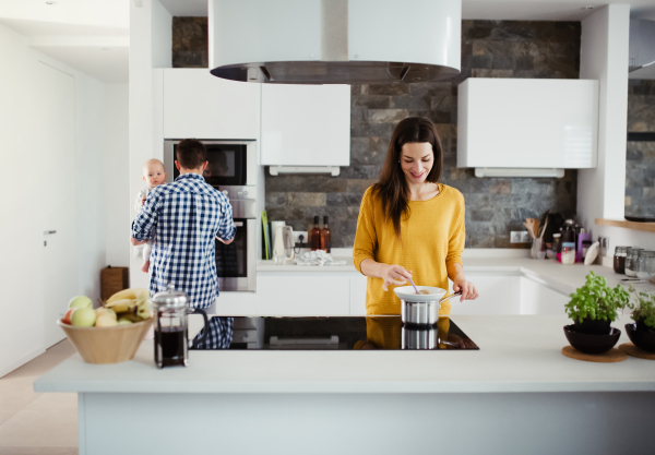 A portrait of young family standing in a kitchen at home, a man holding a baby and a woman cooking.