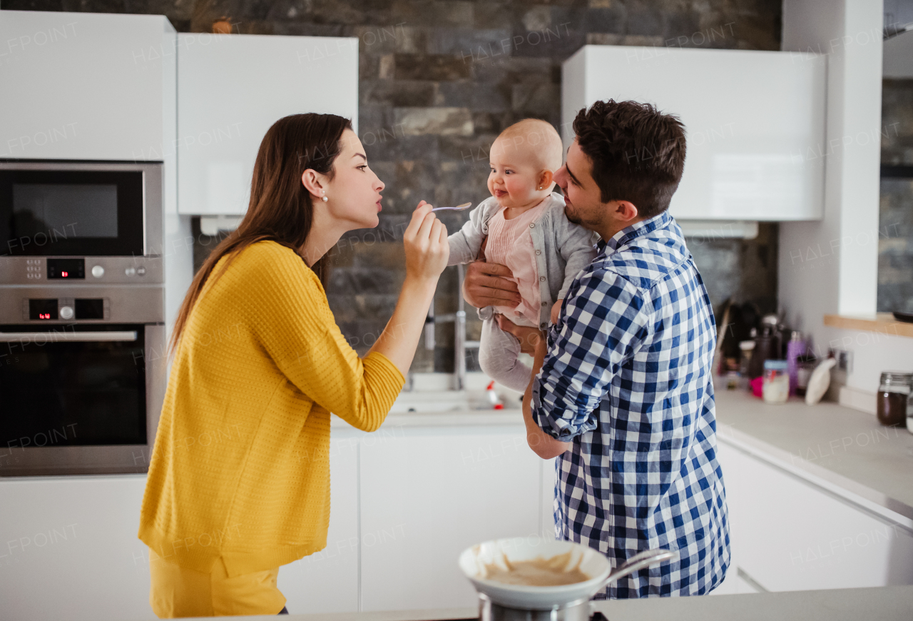 A portrait of young family standing in a kitchen at home, a man holding a baby and a woman feeding her with a spoon.