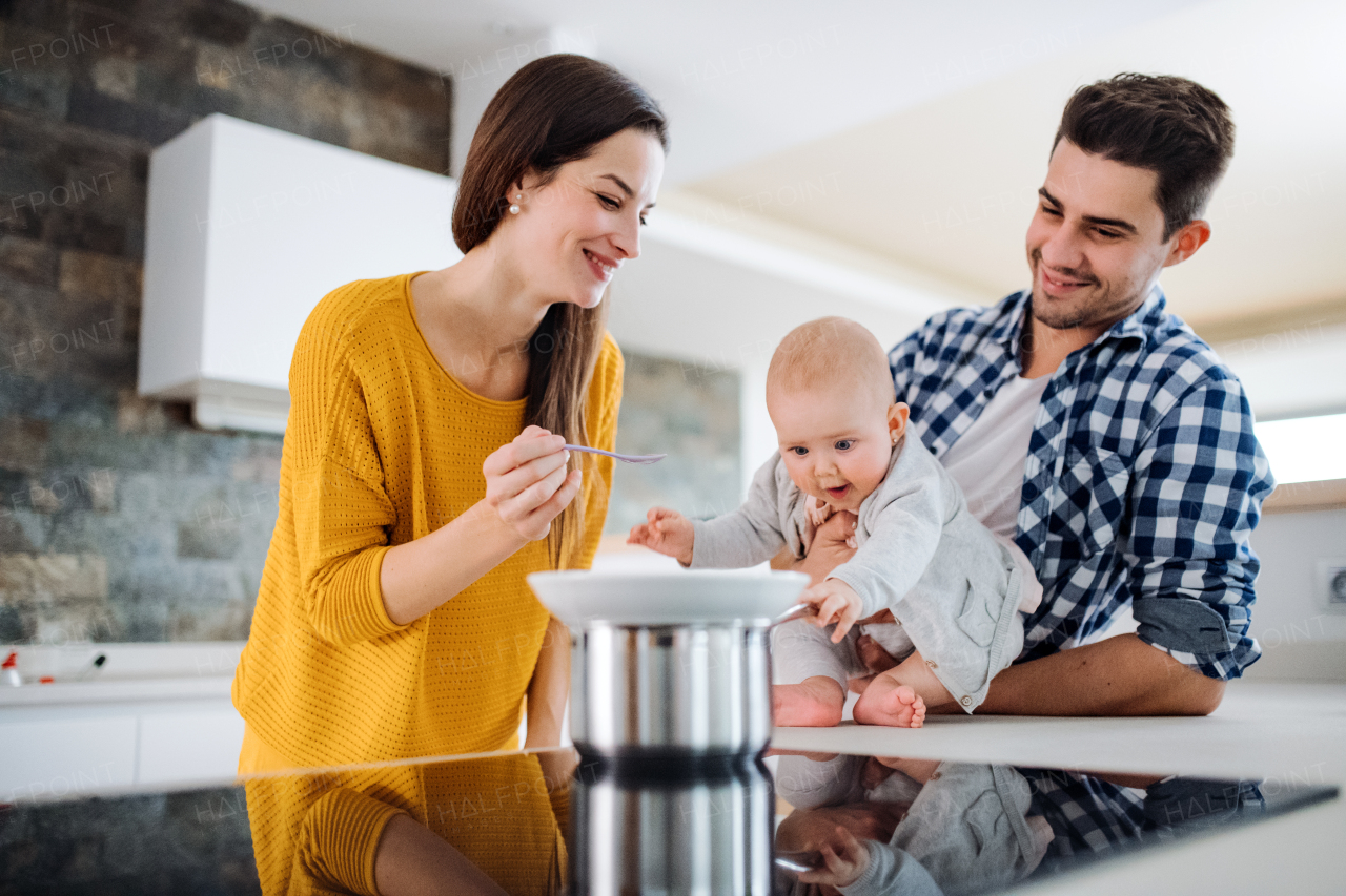 A portrait of young family standing in a kitchen at home, a man holding a baby and a woman feeding her with a spoon.