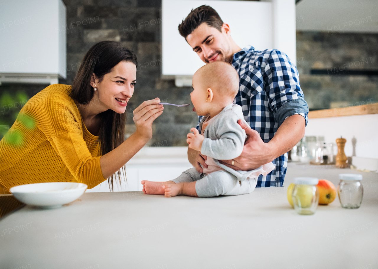 A portrait of young family standing in a kitchen at home, a man holding a baby and a woman feeding her with a spoon.