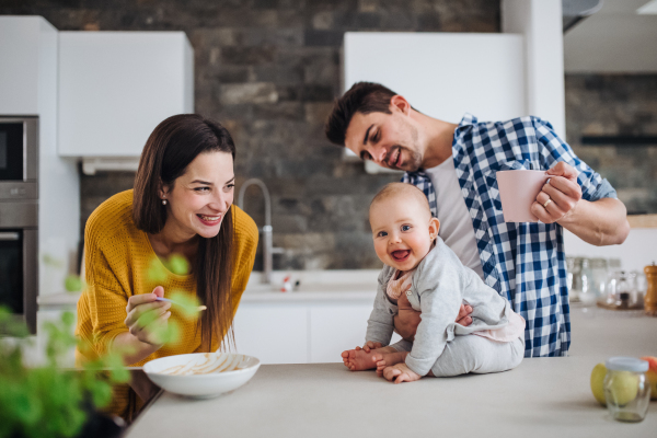 A portrait of young family standing in a kitchen at home, a man holding a baby and a woman feeding her with a spoon.