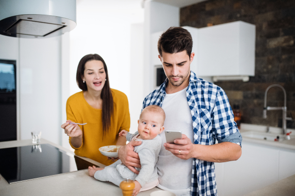 A portrait of young family standing in a kitchen at home, a man with smartphone holding a baby and a woman feeding her with a spoon.