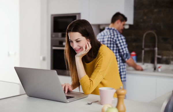 A young attractive woman using laptop in a kitchen and a man washing the dishes.