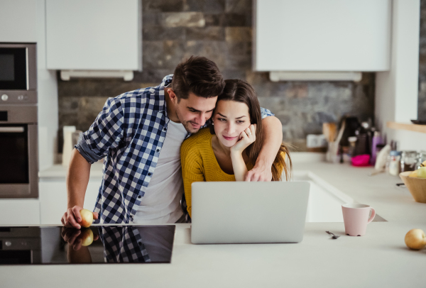 A young happy couple standing in a kitchen at home, using laptop.