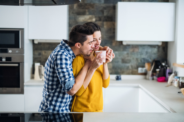 A young happy couple standing in a kitchen at home, drinking coffee.