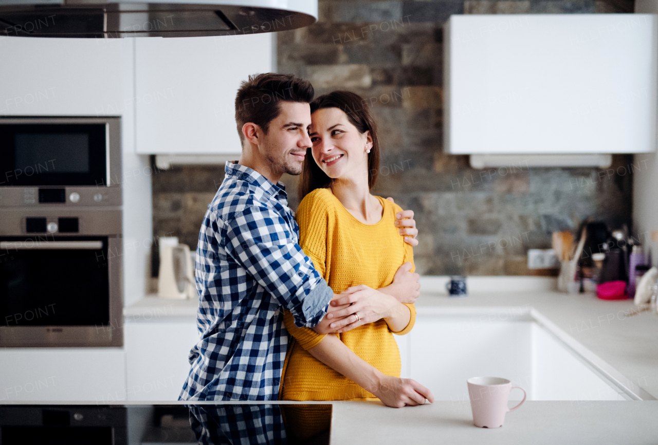 A young happy couple in love standing indoors in a kitchen at home, hugging.