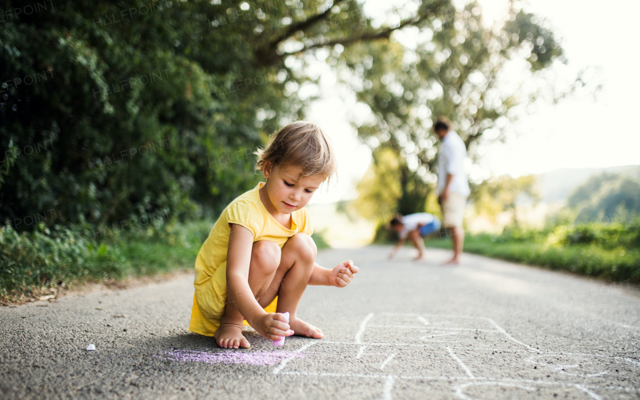 A small cute girl on a road in countryside in sunny summer nature, drawing with chalk.