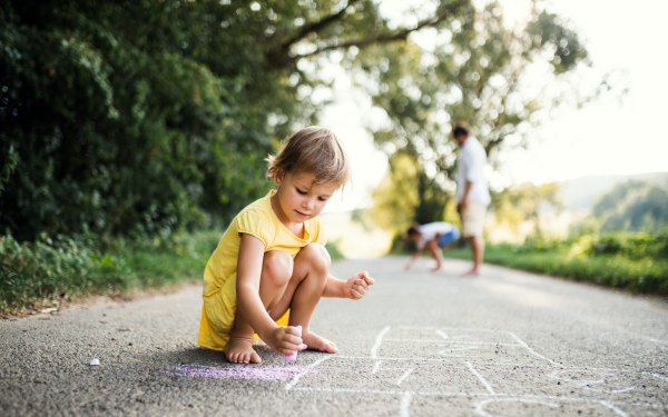 A small cute girl on a road in countryside in sunny summer nature, drawing with chalk.