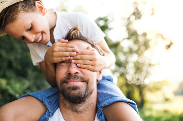 Young father in nature giving a small son a piggyback ride, boy covering man's eyes.