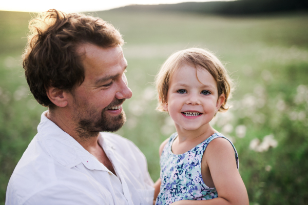 Handsome young father in green sunny summer nature holding his cute small daughter in the arms.