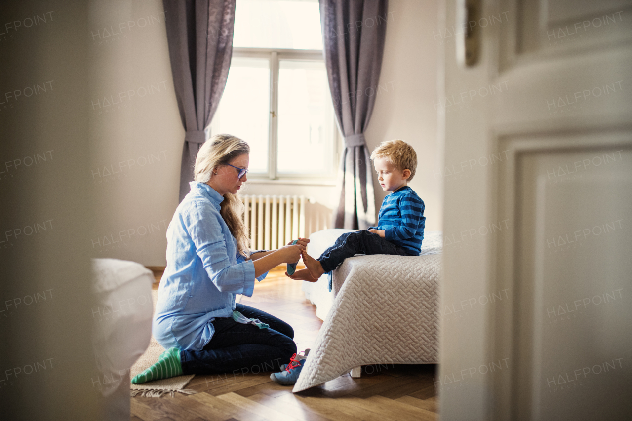 A young mother putting socks on her toddler son inside in a bedroom.