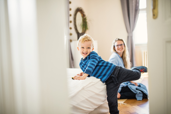 A cheerful toddler boy with young mother in the background inside in a bedroom.