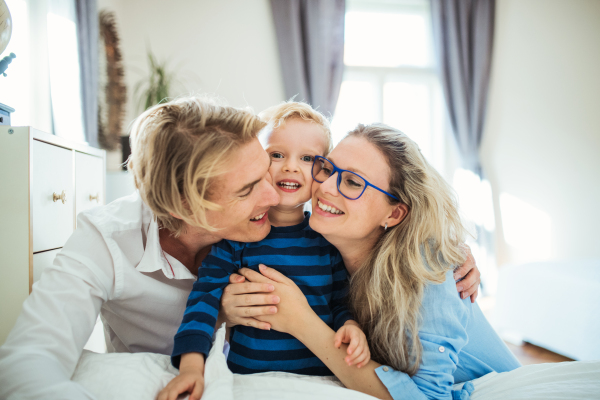 Happy young happy parents hugging their toddler son inside in a bedroom.