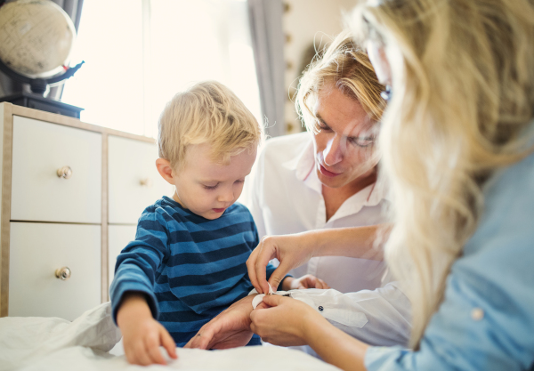 Young parents with their toddler son inside in a bedroom, buttoning a shirt up when dressing.