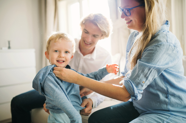 Young happy parents dressing on their toddler son inside in a bedroom.