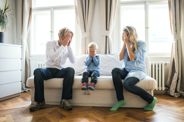 Young parents and toddler son sitting on a sofa inside in a bedroom, covering eyes when playing peekaboo.