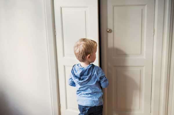 A rear view of toddler boy standing near wardrobe door inside in a bedroom. Copy space.