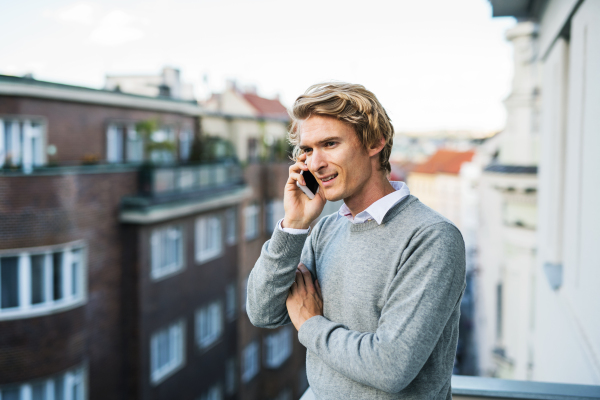 Young man with coffee and smartphone standing on a terrace or balcony in a city, making a phone call.