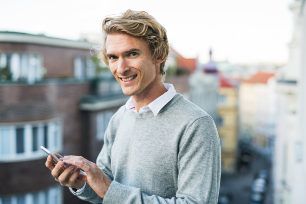 Young man with smartphone standing on a terrace or balcony in a city, texting.