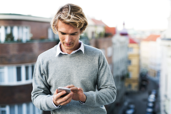 Young man with smartphone standing on a terrace or balcony in a city, text messaging.