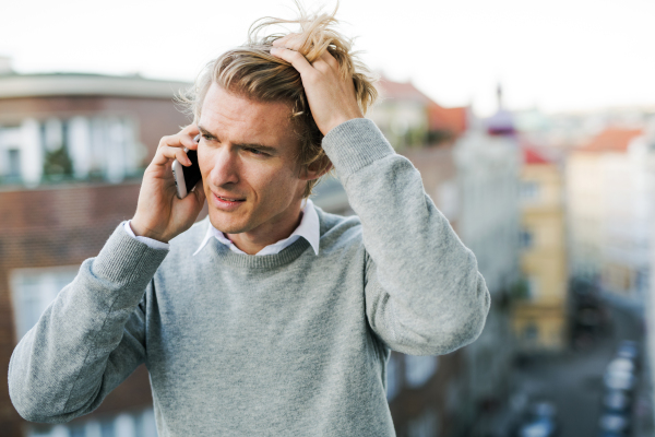 Young man with smartphone standing on a terrace or balcony in a city, making a phone call.