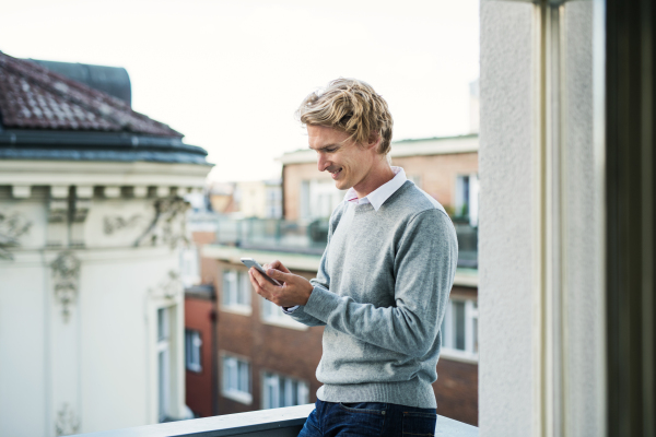 Young man with coffee and smartphone standing on a terrace or balcony in a city, text messaging.