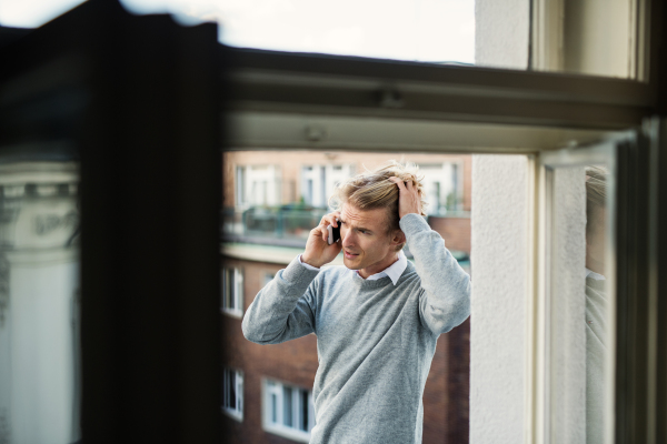 Young man with coffee and smartphone standing on a terrace or balcony in a city, making a phone call.