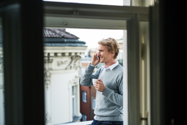 Young man with coffee and smartphone standing on a terrace or balcony in a city, making a phone call.