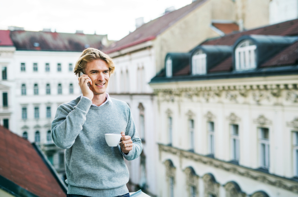 Young man with coffee and smartphone standing on a terrace or balcony in a city, making a phone call.