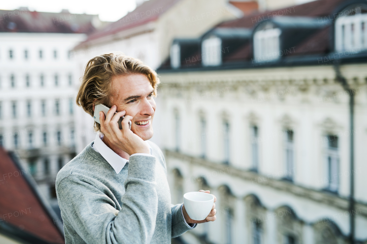 Young man with coffee and smartphone standing on a terrace or balcony in a city, making a phone call. Copy space.