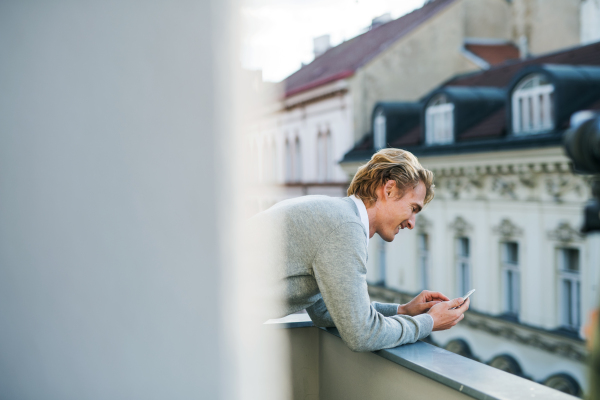 Young man with smartphone standing on a terrace or balcony in a city, texting. Copy space.