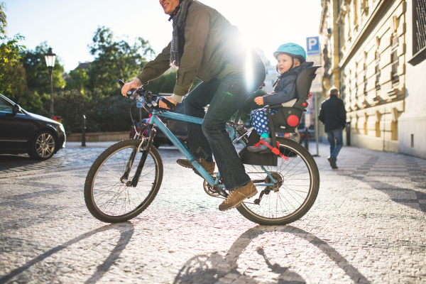 A small toddler boy with helmet sitting in bicycle seat with unrecognizable father outdoors on a street in city.