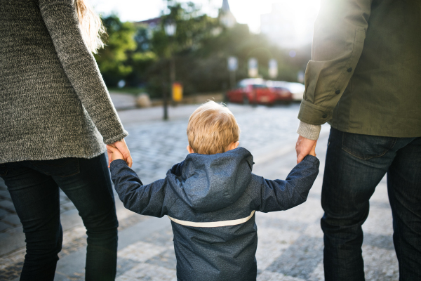 A rear view of small toddler boy with unrecognizable parents walking outdoors in city, holding hands.