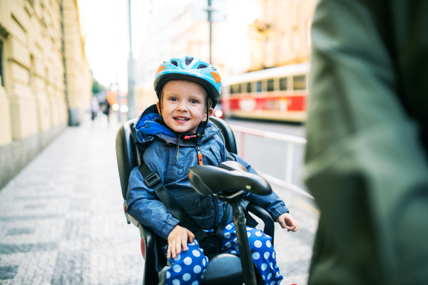 A small toddler boy with helmet sitting in bicycle seat with unrecognizable father outdoors on a street in city.