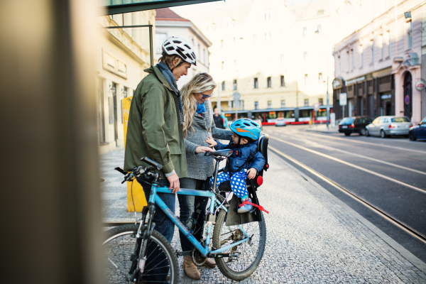 A small toddler boy with helmet sitting in bicycle seat with young parents outdoors on a street in city.