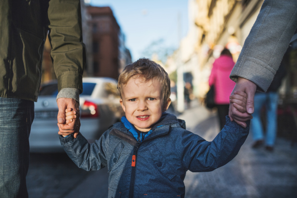 A small toddler boy with unrecognizable parents walking outdoors in city, holding hands.