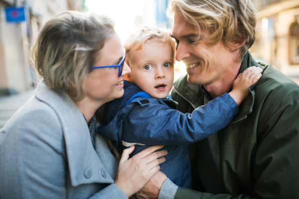 Young happy parents hugging their toddler son outdoors in city.