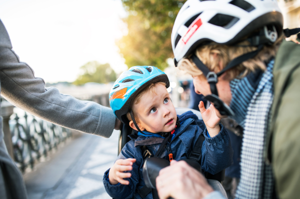 A small toddler boy with bicycle helmet and unrecognizable young parents outdoors in city.