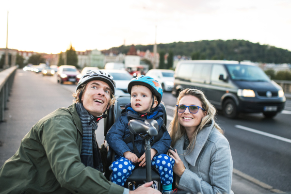 A small toddler boy with helmet sitting in bicycle seat with young parents outdoors on a street in city.