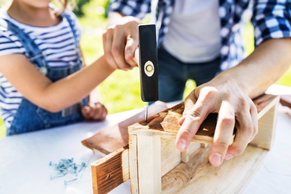 Unrecognizable father with a small daughter outside, making wooden birdhouse or bird feeder.