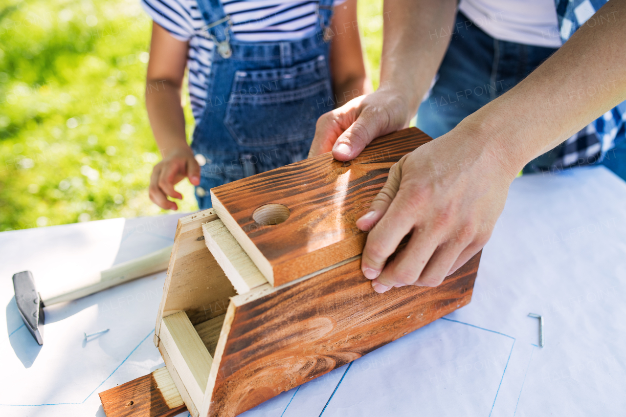 Unrecognizable father with a small daughter outside, making wooden birdhouse or bird feeder.