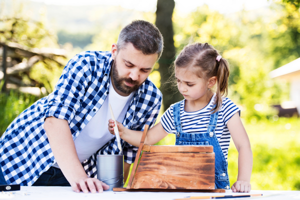 Mature father with a small daughter outside, making wooden birdhouse or bird feeder.