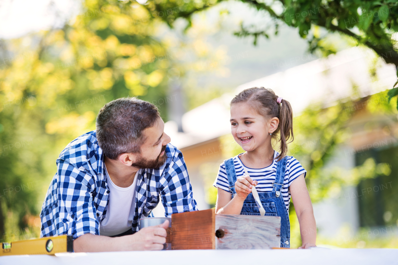 Mature father with a small daughter outside, painting. Wooden birdhouse or bird feeder making.