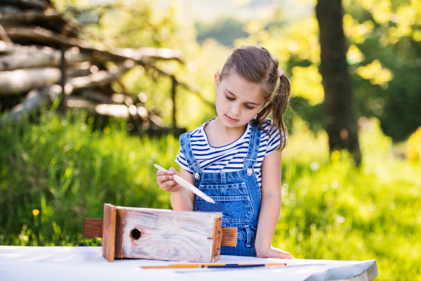 A happy small girl with a paintbrush outside, painting a wooden birdhouse or bird feeder.