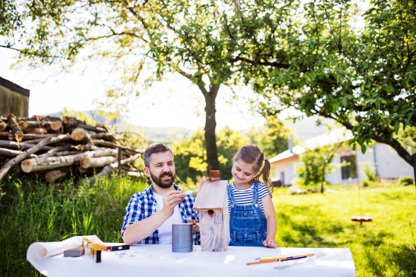 Mature father with a small daughter outside, painting. Wooden birdhouse or bird feeder making.